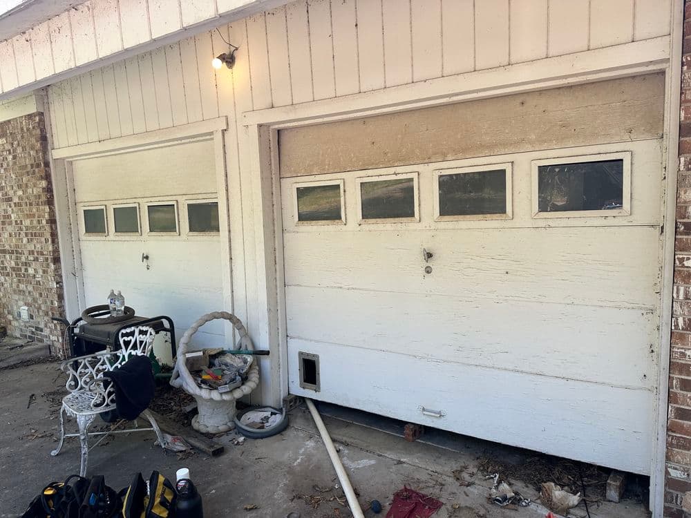 Two wooden garage doors with windows, surrounded by scattered tools and a chair.