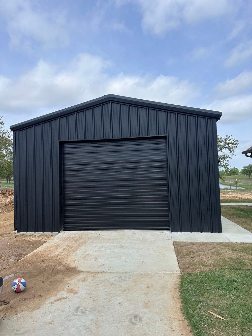 Black metal garage with roll-up door, concrete driveway, and grassy area in front.