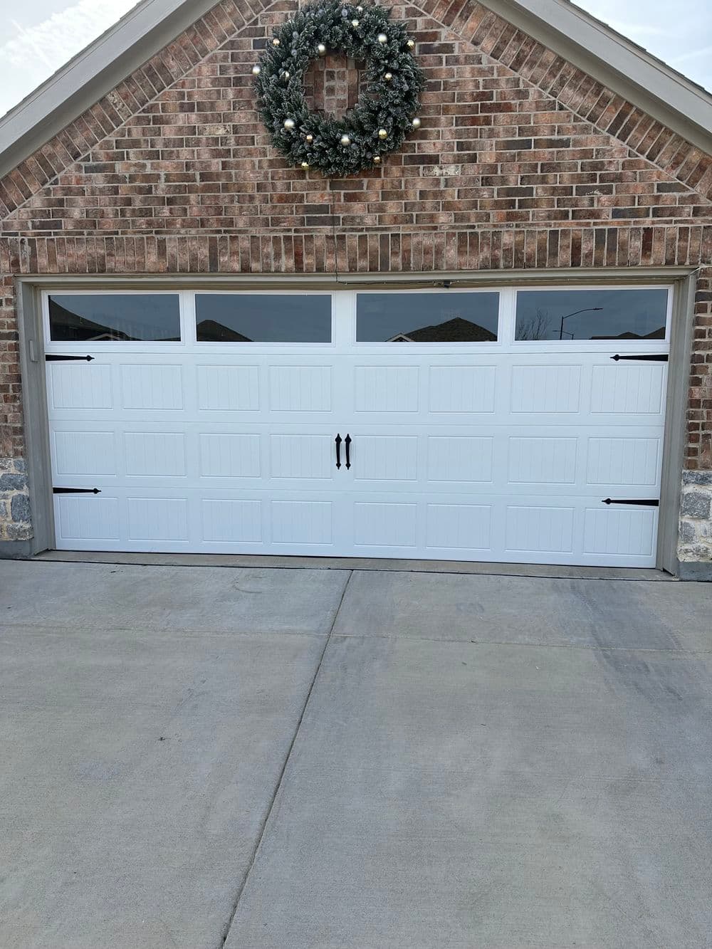 White garage door with black handles, decorated with a wreath, set against a brick wall.