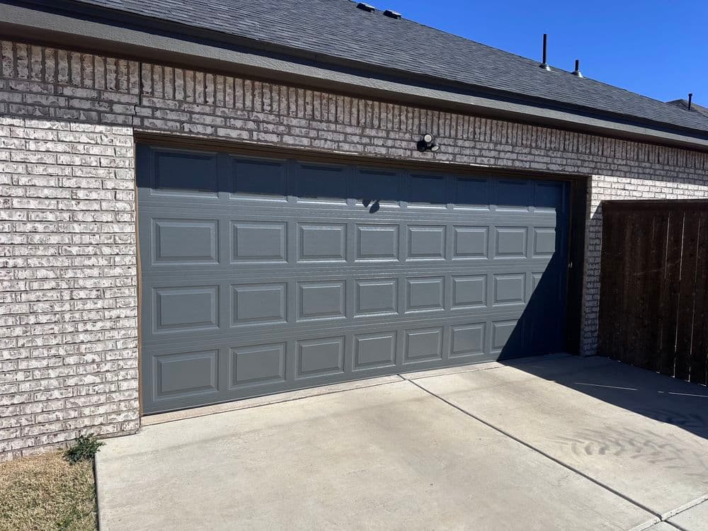 Gray garage door on a brick house with a concrete driveway and blue sky backdrop.